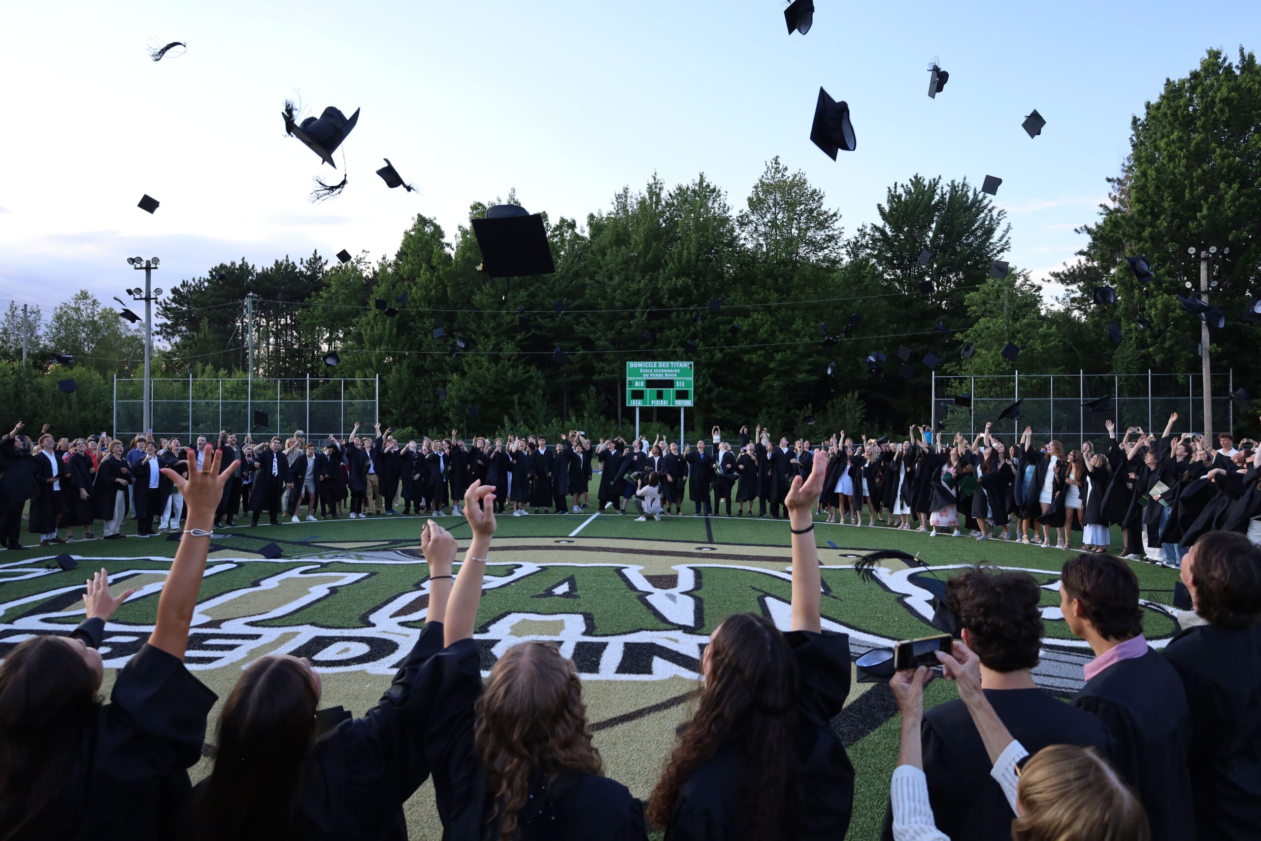 Un grand groupe de diplômés en toge noire lance joyeusement ses mortiers dans les airs lors d'une cérémonie de graduation en plein air. L'événement se déroule sur un terrain de sport extérieur verdoyant, entouré de grands arbres sous un ciel de fin de journée. Au premier plan, on voit de dos les têtes et les bras levés de plusieurs étudiants qui immortalisent le moment, tandis qu'en arrière-plan, une longue rangée d'étudiants célèbre devant un tableau indicateur vert.